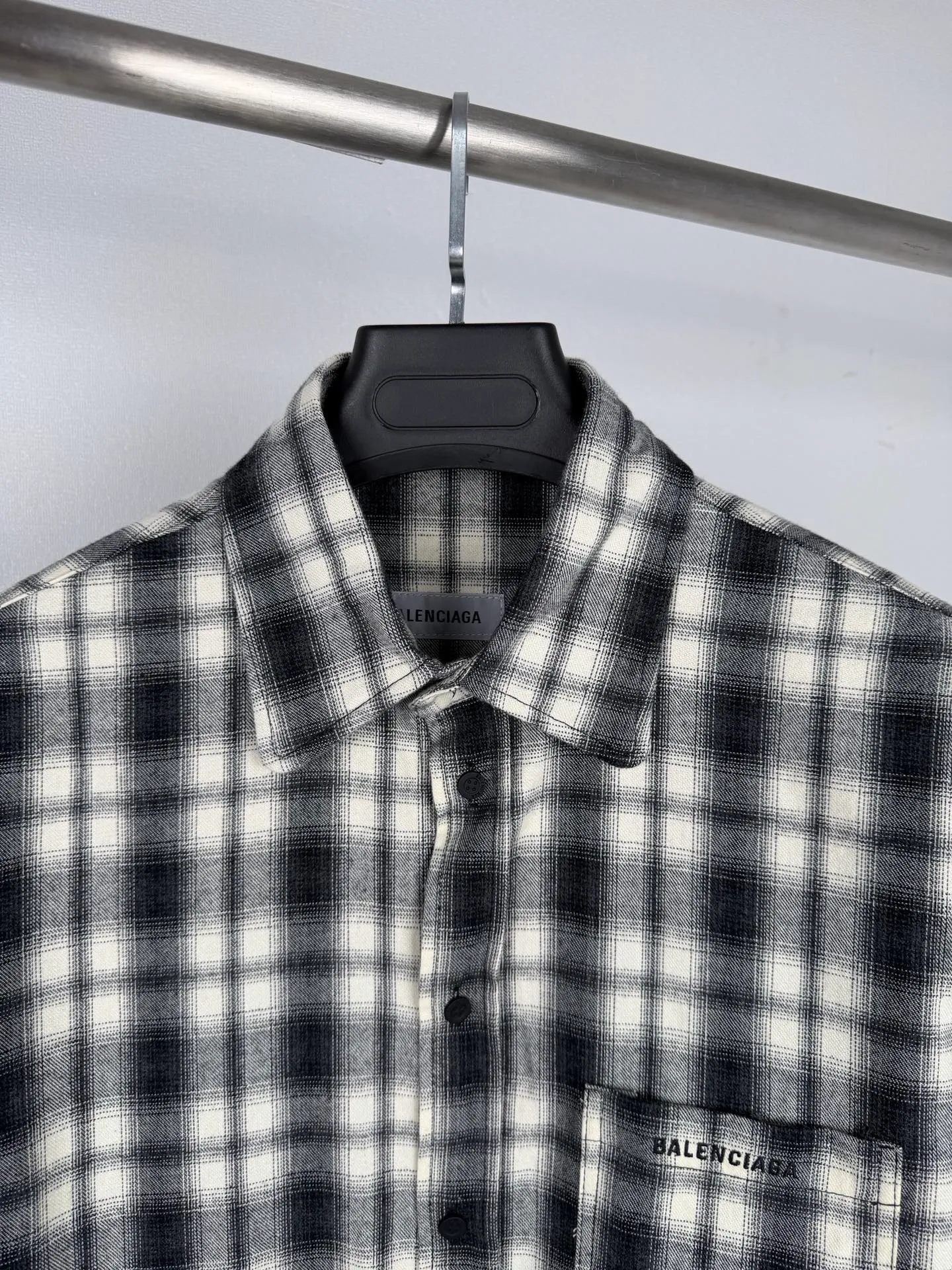 BALENCIAGA/balenciaga Black White plaid pocket embroidered logo shirt jacket The technical difficulties of this plaid shirt focus on fabric finishing, plaid matching and component sewing details. The fabric has been sanded and distressed to create a fine velvet feel on the surface, changing the original luster of the fabric and making the Black and White plaid colors transition softly. At the same time, the drape properties of the fabric itself are retained, taking into account the looseness of the upper body and the silhouette of the body, without appearing soft and shapeless. The biggest test of plaid garments comes from the alignment of the pieces. The front and rear body, large and small sleeves, and chest pockets are all independent pieces. In the cutting stage, the horizontal and vertical axes of the plaid need to be aligned, and the external chest bag is cut separately. The plaid on the bag surface needs to form a visual coordination with the main body of the garment. Cutting and typesetting will produce additional fabric loss, and there are high requirements for the cutting accuracy of the cutting bed typesetting and workers. The collar adopts a double-layer combined structure, and the collar surface and collar seat are double-stitched with even and regular stitching spacing to ensure the lapel's long-lasting shape and resistance to warping and deformation after repeated bending. The placket is sewn with regular topstitching and paired with Black matte buttons. The button points need to be aligned with the checkered lines to avoid damaging the overall visual order. The splicing of the cuffs also takes into account the grain direction, and the overlocking process reinforces the seams to improve the durability of the garment. After the entire garment is sewn, it also undergoes a garment washing process to further strengthen the Retro texture of the fabric, unify the tone and texture of the entire garment, weaken the harshness of industrial production, and finally present a relaxed Retro garment effect. Size: 46 48 50 52
